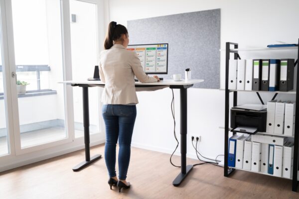 Kobieta pracuje przy komputerze stojąc przy biurku regulowanym elektrycznie w nowoczesnym biurze.